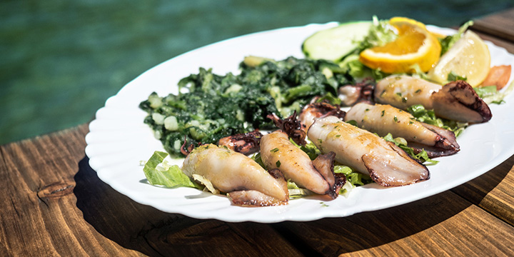 Grilled squid with boiled potatoes and chard (blitva) on white plate with sea on background. Traditional Croatian food. Sunny day.