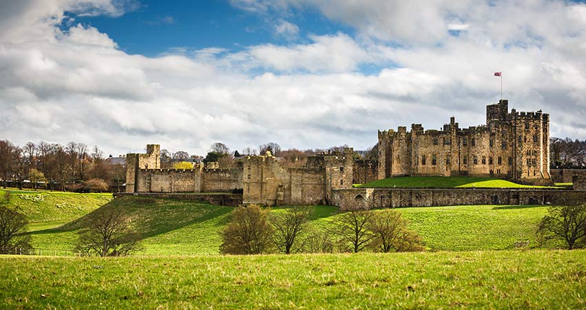 Alnwick Castle in England on a bright day, surrounded by green fields and scattered trees.
