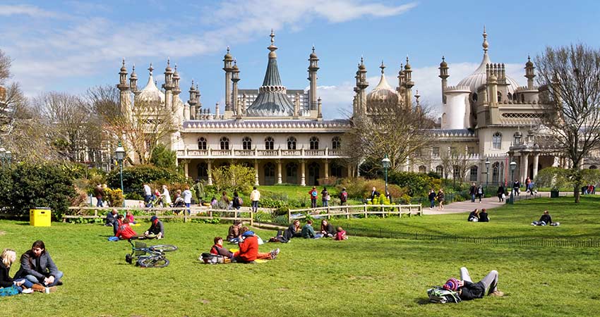 People relaxing on the grass in front of the Royal Pavilion in Brighton, England, under a blue sky.