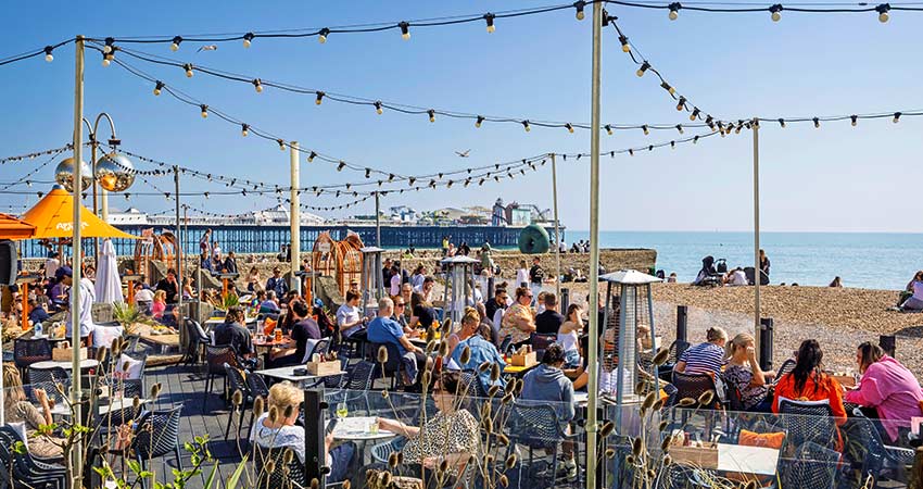 People dining at a lively beachside restaurant in Brighton, England, with a view of the pier and sea.