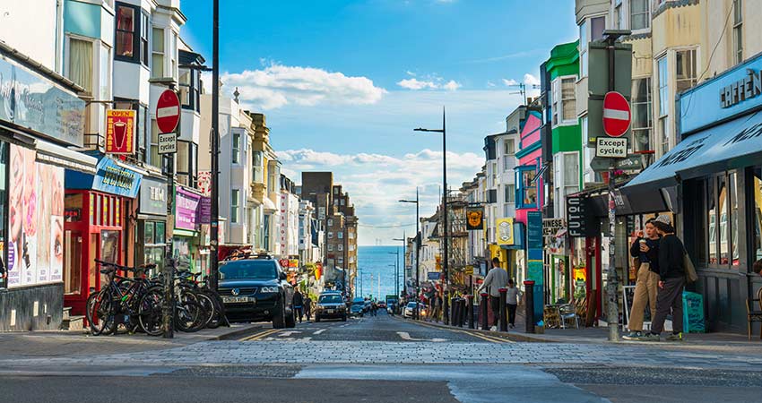 Colorful shops and cafés line a lively street in Brighton, England, with a view of the sea in the distance.