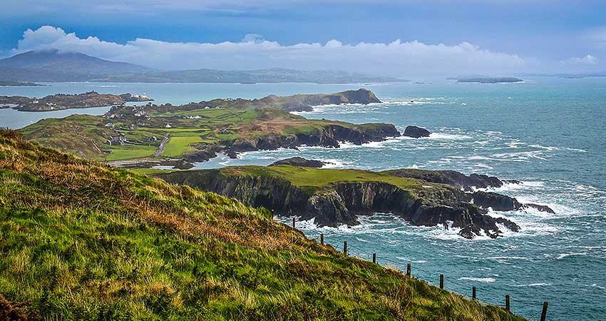 Dramatic coastal cliffs and rolling green fields along Brow Head, County Cork, Ireland, with the Atlantic Ocean crashing below.