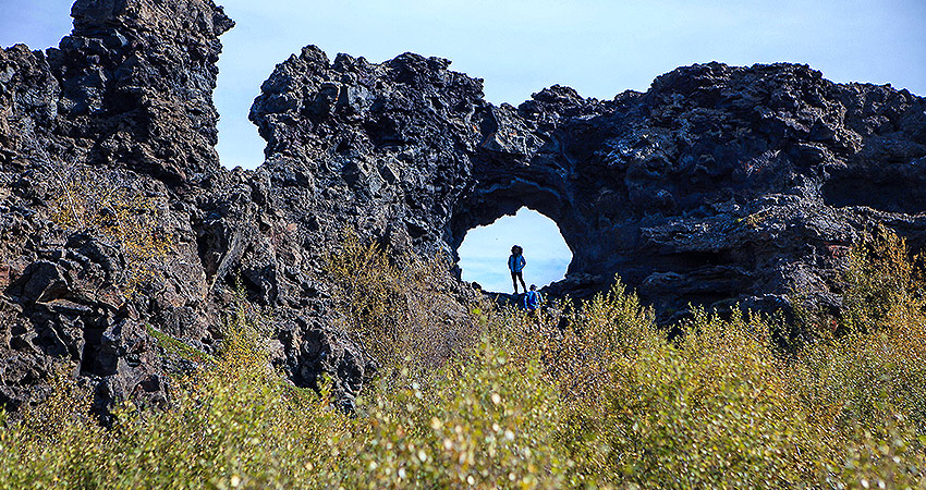 Hikers standing inside a natural lava arch at Dimmuborgir, Iceland, surrounded by volcanic rock formations and shrubs.