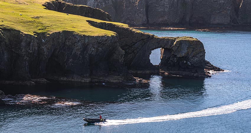 Small boat passing by a dramatic sea arch and cliffs on Fair Isle, Scotland, with clear blue water and grassy headlands.