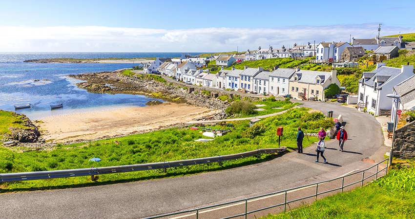 Coastal village on the Isle of Islay, Scotland, with whitewashed cottages, a small beach, and people walking along a winding seaside road.