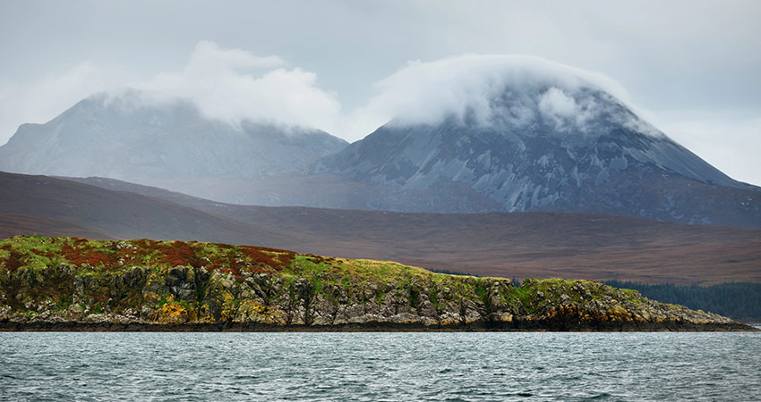 Misty mountains on the Isle of Jura, Scotland, with low clouds covering the peaks and a rugged, mossy shoreline in the foreground.