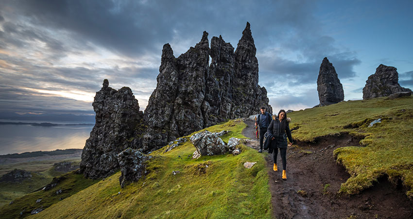 Two hikers walking along a grassy trail beneath dramatic rock formations on the Isle of Skye, Scotland, during a cloudy sunset.