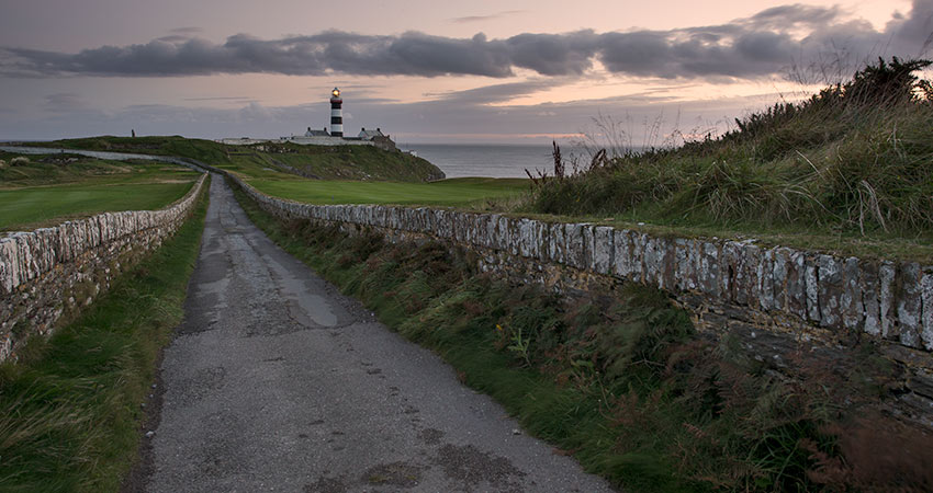 Kinsale Lighthouse at sunset, viewed from a narrow road lined with stone walls and grassy banks along the Irish coast.