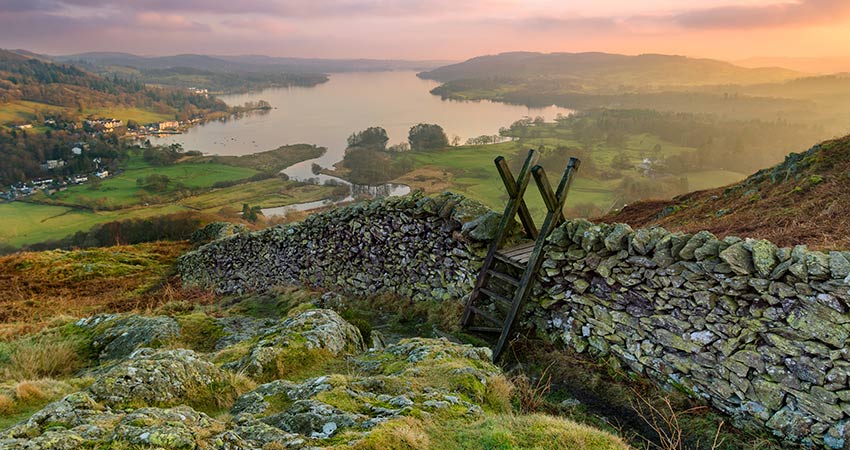 Stone stile crossing a dry stone wall overlooking Lake Windermere and the surrounding countryside at sunset in England’s Lake District.
