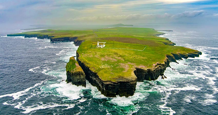 Aerial view of Loop Head, Ireland, with dramatic cliffs, crashing waves, and a white lighthouse perched near the edge of the grassy peninsula.