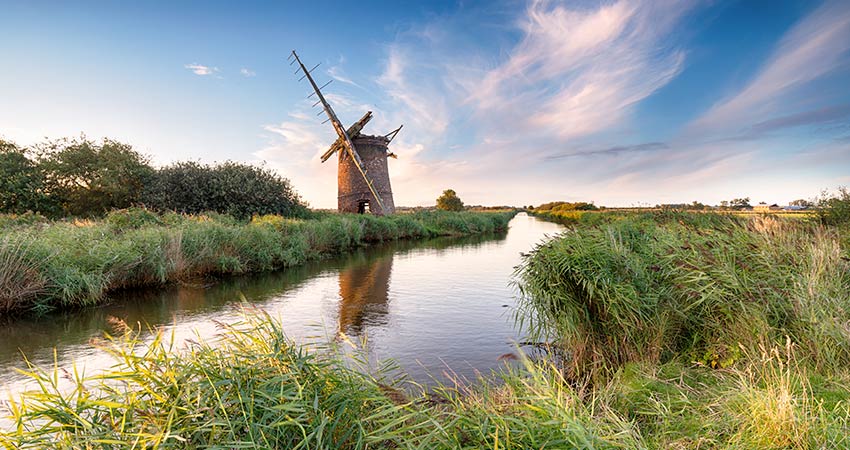 Historic windmill beside a tranquil waterway in the Norfolk Broads at sunset.