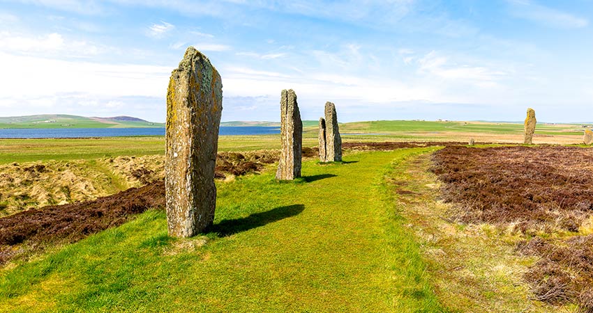 Ancient standing stones on a grassy path in the Orkney Islands, Scotland, with open moorland and coastal views in the background.