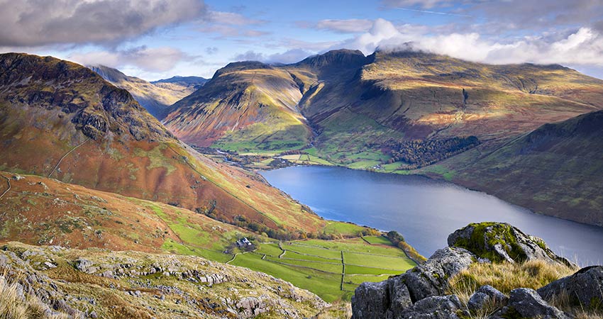 View of Scafell Pike and surrounding peaks in England’s Lake District, with a deep blue lake and patchwork farmland in the valley below.