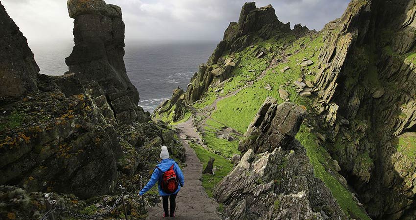 Hiker descending stone steps on Skellig Michael, surrounded by dramatic cliffs and lush green slopes overlooking the Atlantic Ocean.