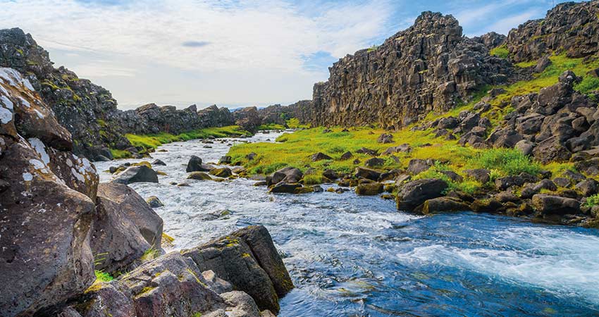 A clear stream flows through rocky cliffs and green moss at Thingvellir National Park in Iceland.