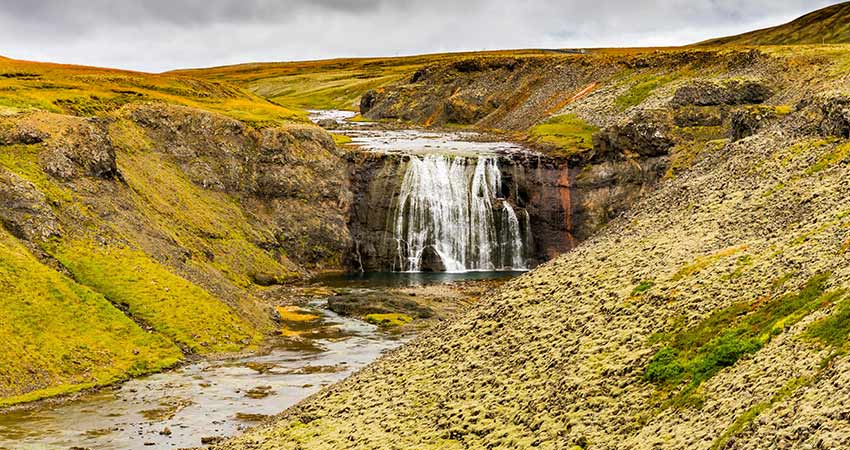 Þórufoss waterfall in Iceland cascading over rocky cliffs into a calm pool, surrounded by mossy hills.