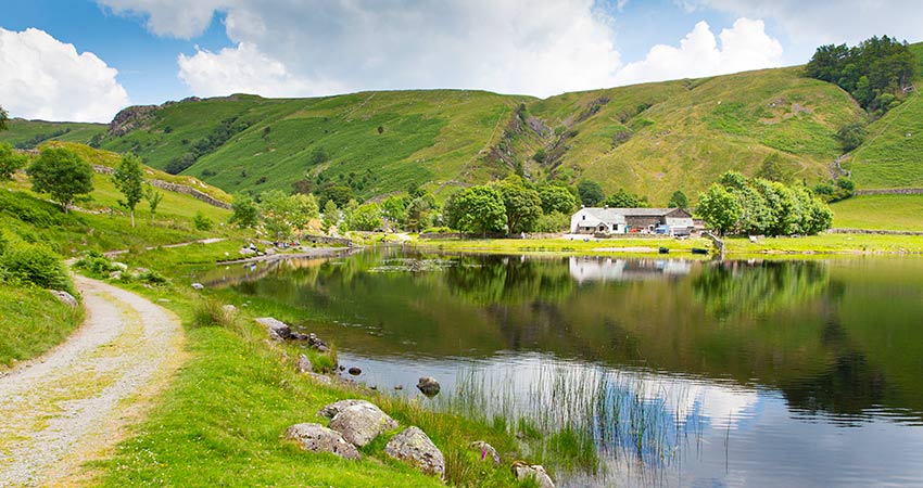 Peaceful scene of Watendlath Tarn in the Lake District, England, with a walking path, still water, green hills, and a white farmhouse in the distance.